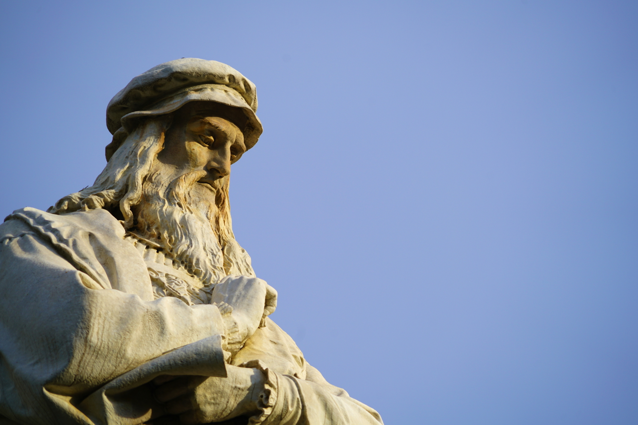 Kopf der Leonardo da Vinci-Statue in Mailand Steinfigur eines nachdenklichen Mannes mit Bart und Mütze vor blauem Himmel.