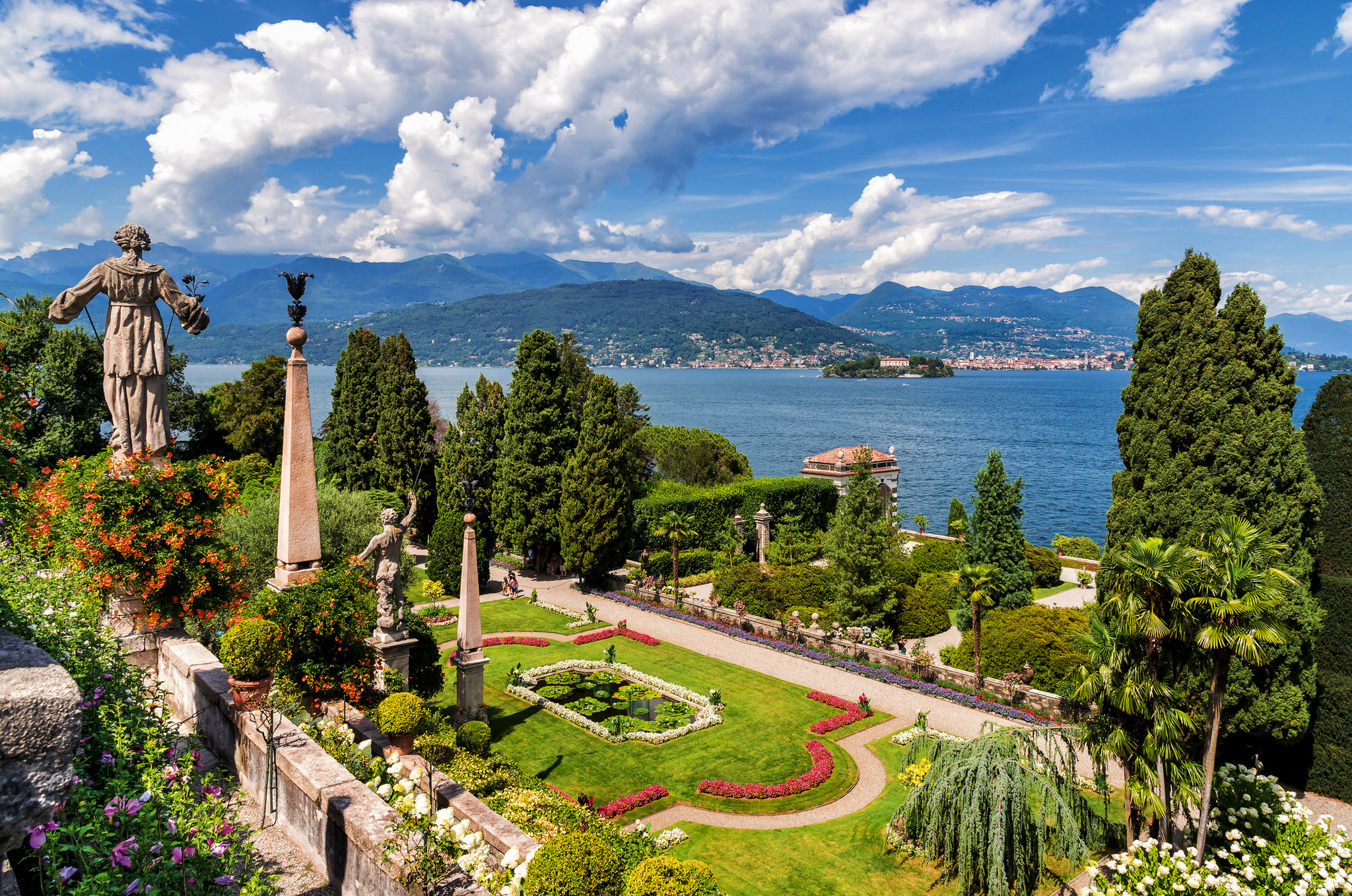 Gartenanlage mit Statuen, Bäumen und Blick auf einen See und Berge im Hintergrund.