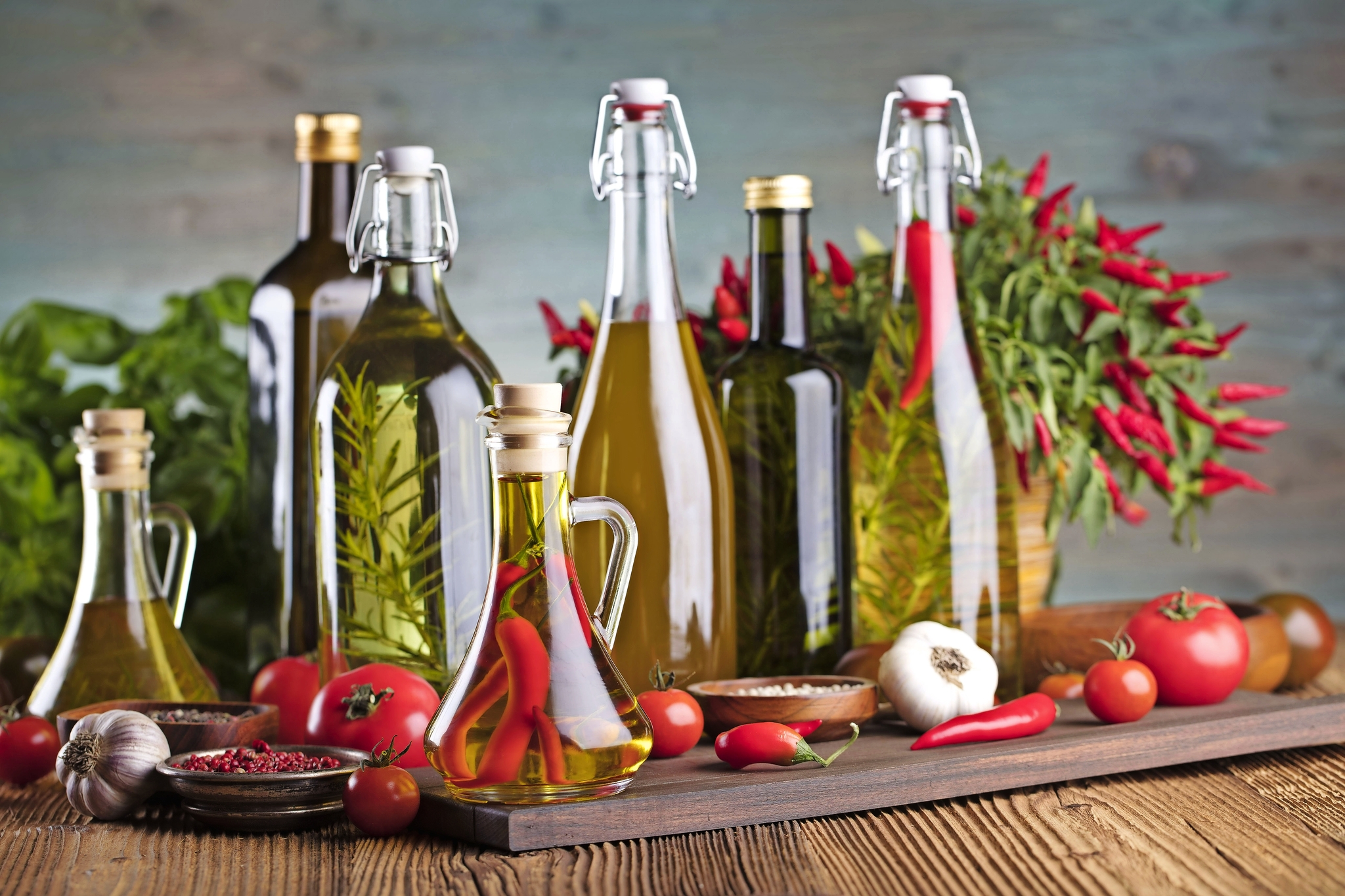 Verschiedene Flaschen mit Olivenöl, Kräutern, Paprika und Gemüse auf Holzuntergrund.
