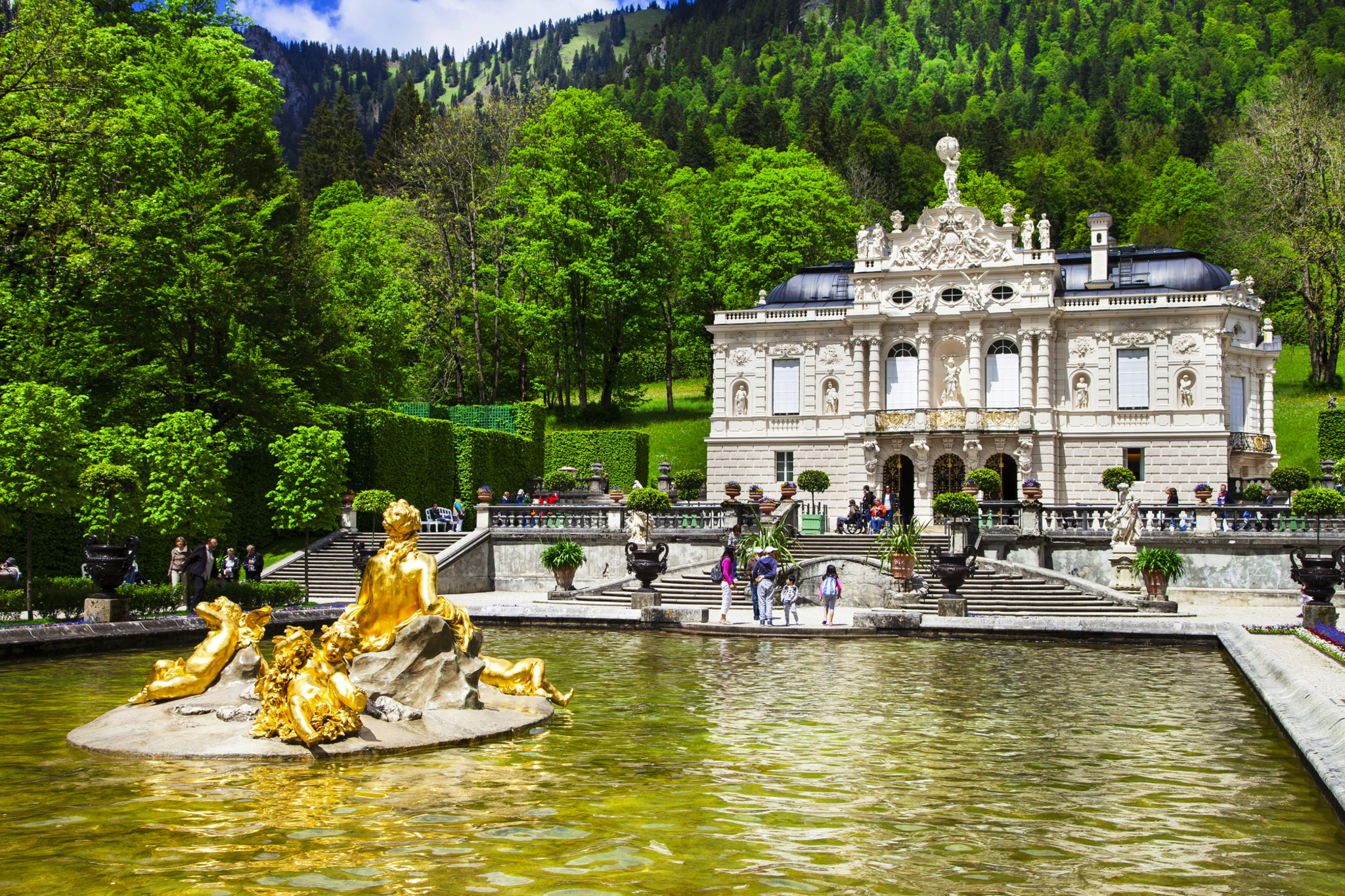 Schloss mit vergoldetem Brunnen im Park, umgeben von Wald und Grünflächen.
