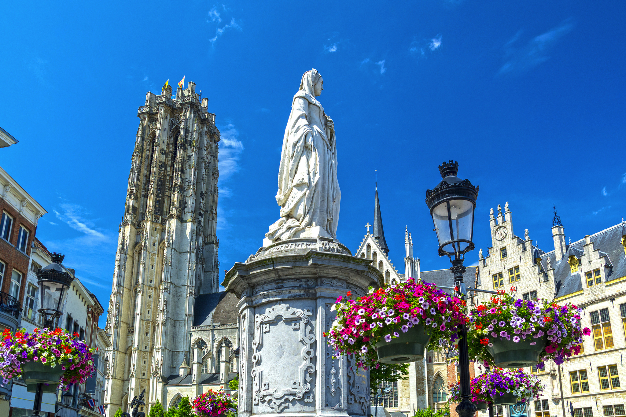 Mechelen in der Provinz Antwerpen in Belgien Statue mit Glockenturm im Hintergrund und blühenden Blumen im Vordergrund.