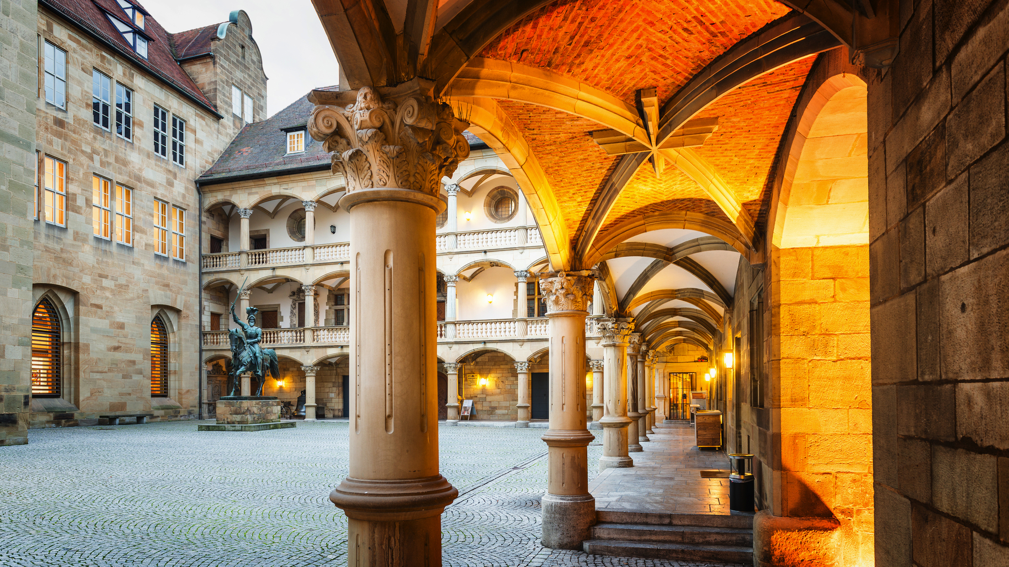 Historischer Innenhof mit Arkaden und Statue, abends beleuchtet.