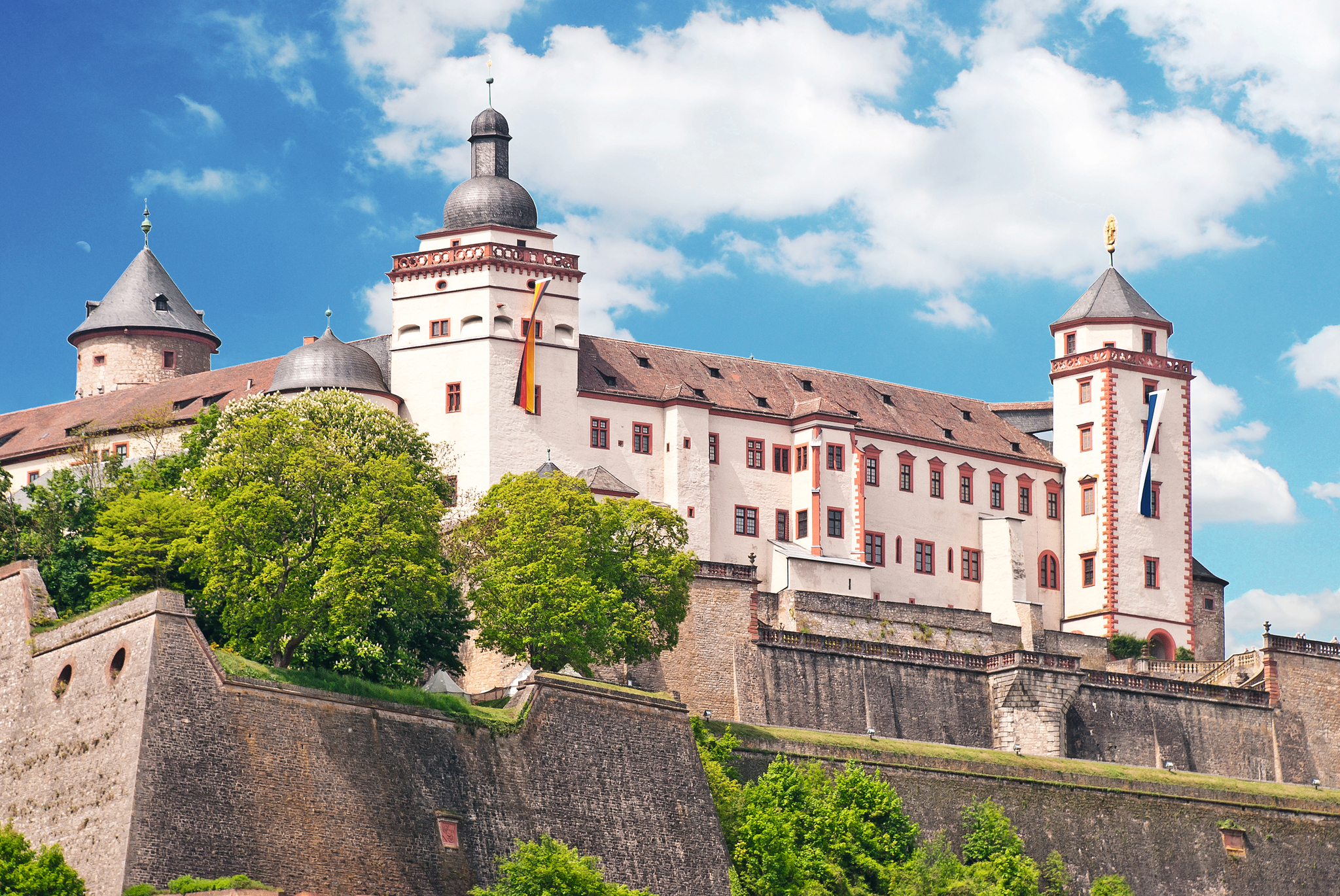 Burg auf Hügel mit Türmen und Bäumen, blauer Himmel im Hintergrund.