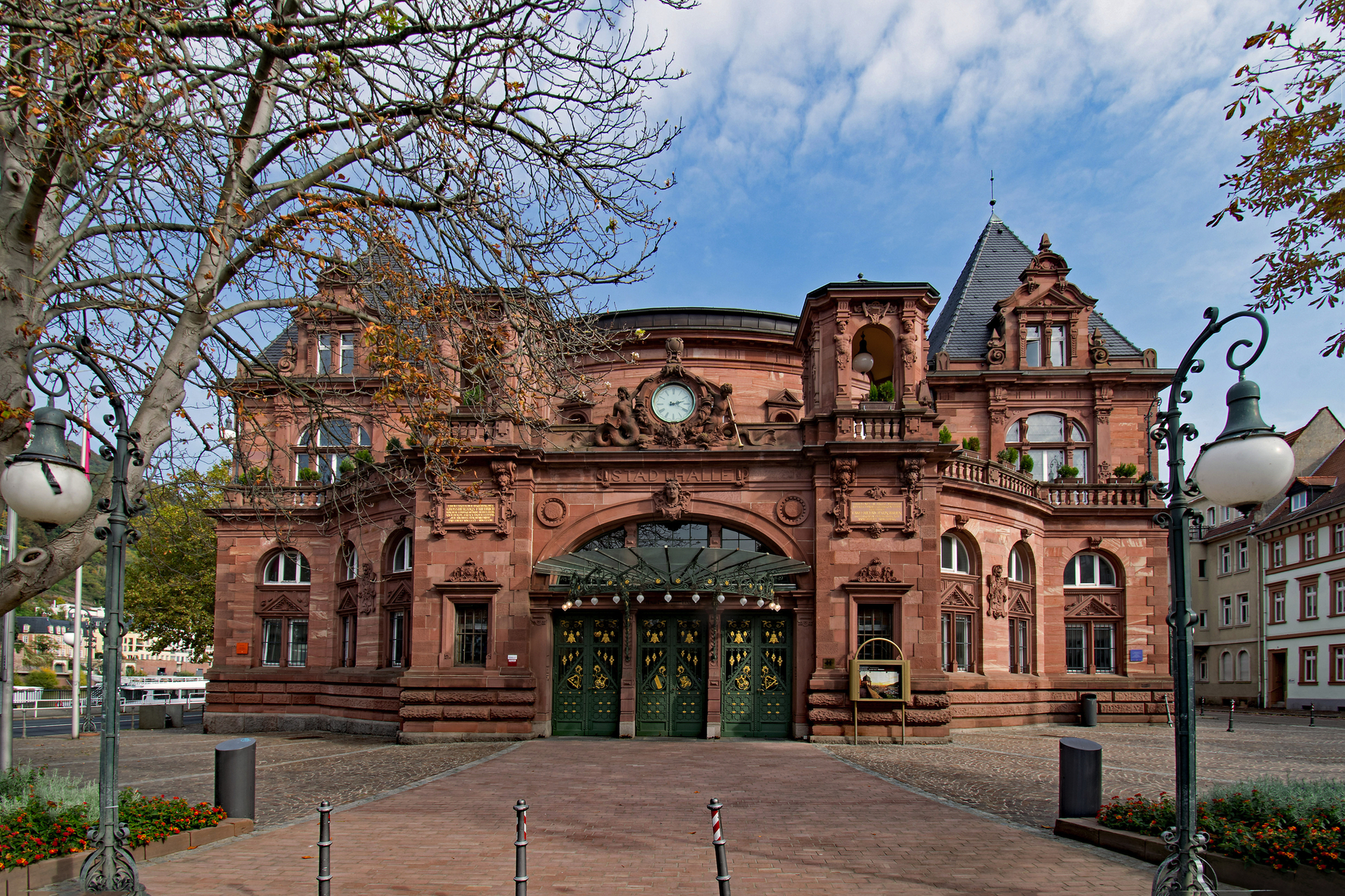 alte Stadthalle von Heidelberg Alte Stadthalle in Heidelberg, Deutschland, umgeben von herbstlich verfärbten Bäumen, im Vordergrund gepflasterter Platz, historisches Gebäude mit roter Fassade und dekorativen Elementen, blauer Himmel im Hintergrund.