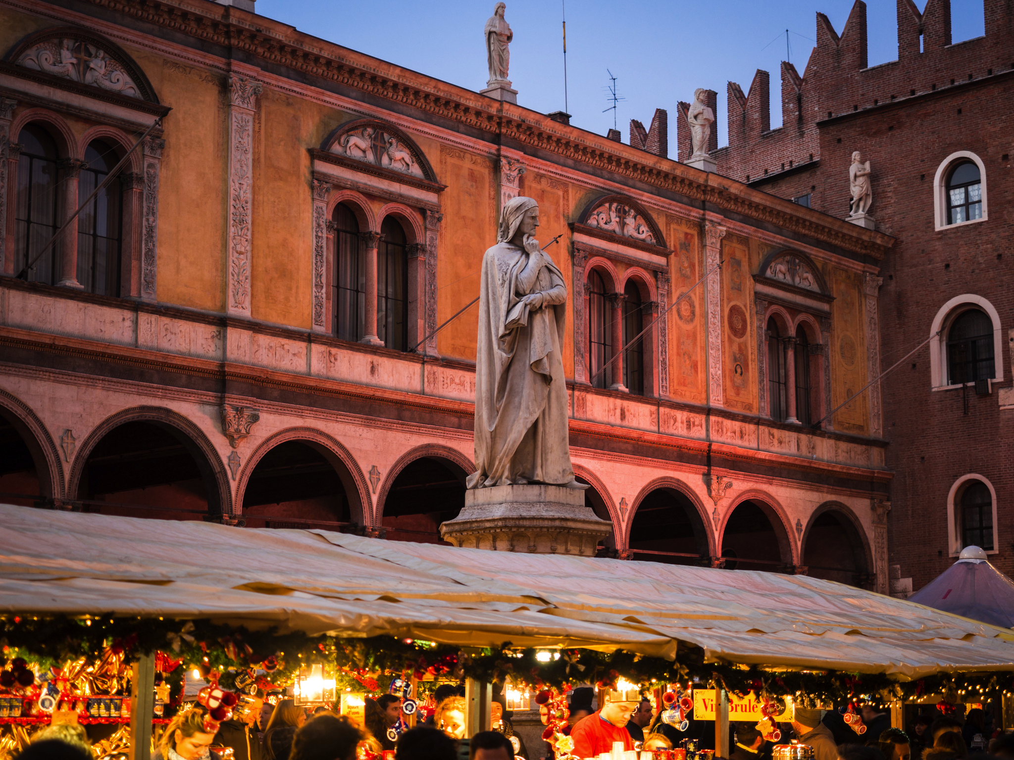 Statue von Dante Alighieri auf der Piazza dei Signori während des Christkindmarkts Statue von Dante Alighieri auf der Piazza dei Signori in Verona, umgeben von festlicher Weihnachtsmarktdekoration während der Adventszeit.
