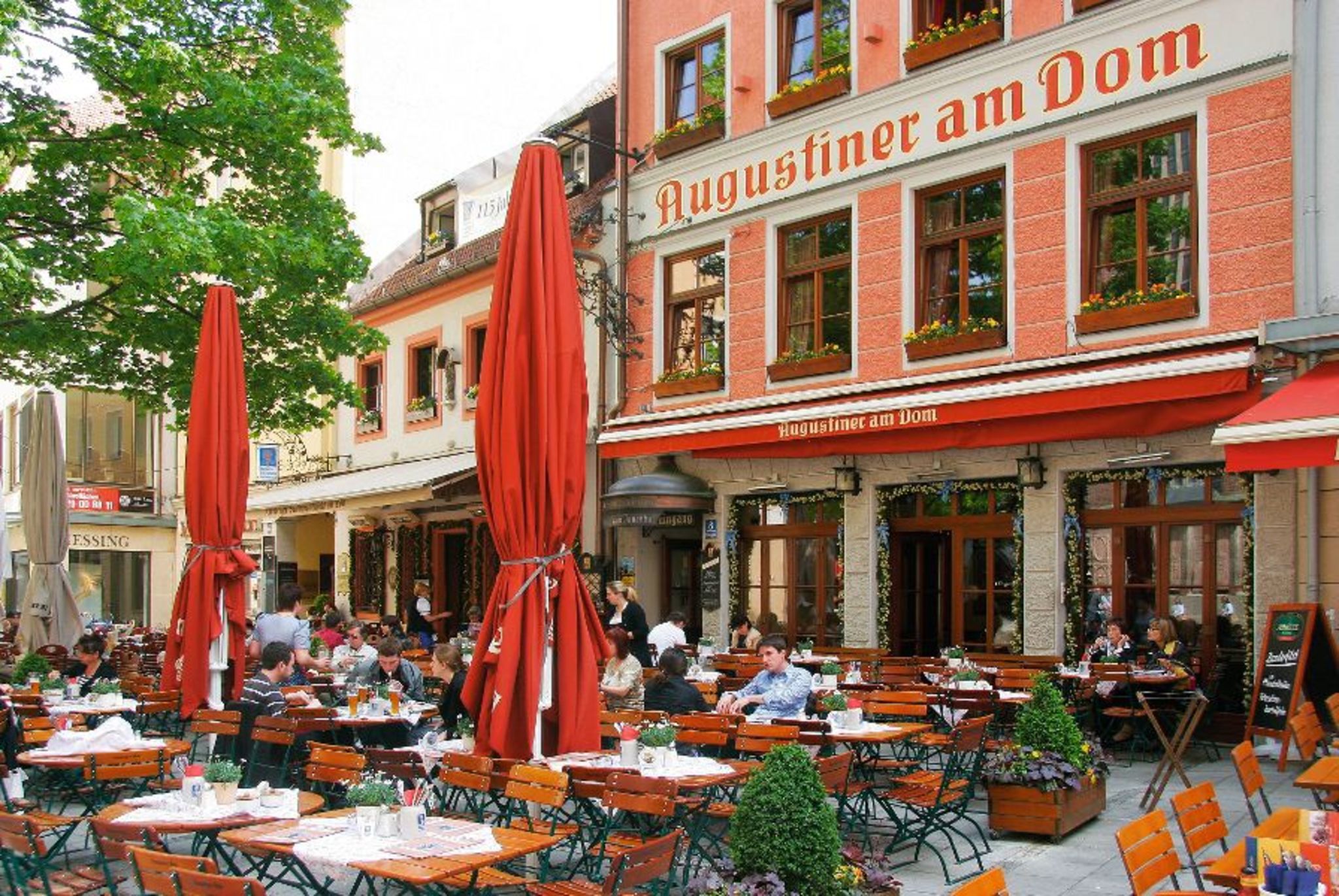 Außenbereich des Restaurants Augustiner am Dom mit Gästen an Tischen und roten Sonnenschirmen.