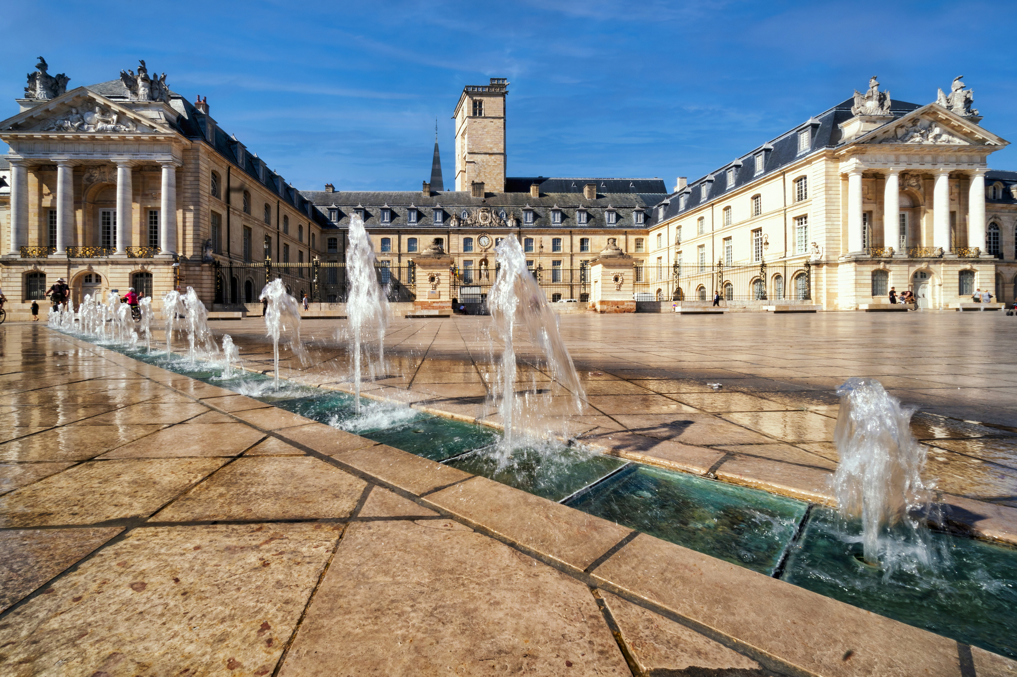 Platz mit Brunnen vor historischem Gebäude bei sonnigem Wetter.