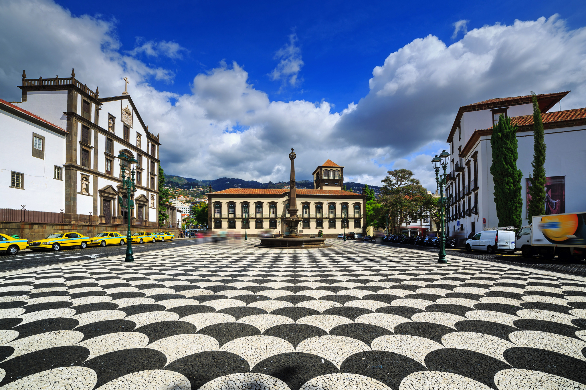 Stadtplatz Praca do Municipio in Funchal, Madeira, Portugal mit gepflastertem Bodenmuster, historischen Gebäuden, einer zentralen Statue und umgeben von geparkten Autos bei leicht bewölktem Himmel.