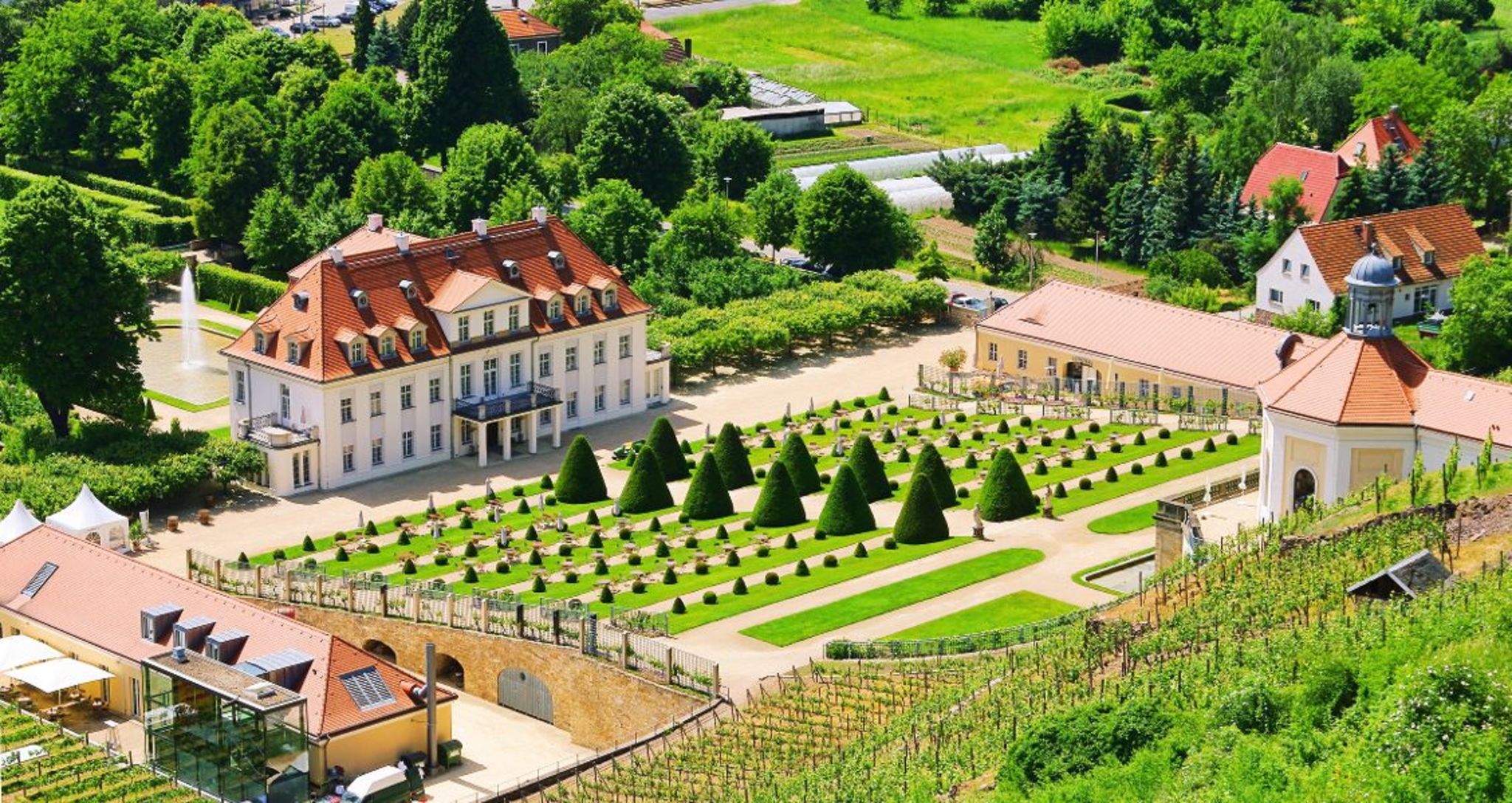 Schloss mit symmetrischem Garten und Weinbergen in grüner Landschaft aus der Vogelperspektive.