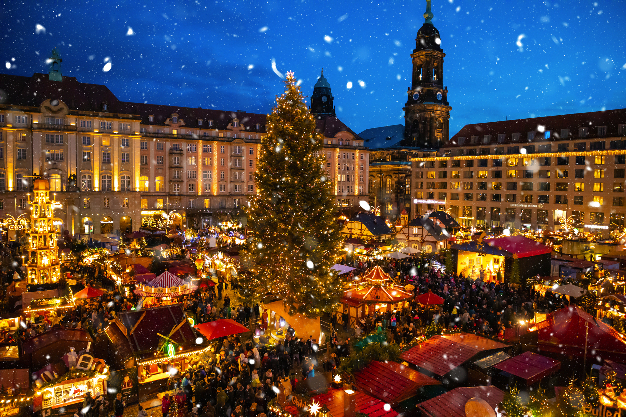 Weihnachtsmarkt am Abend mit Lichtern und großem geschmücktem Tannenbaum im Schnee.