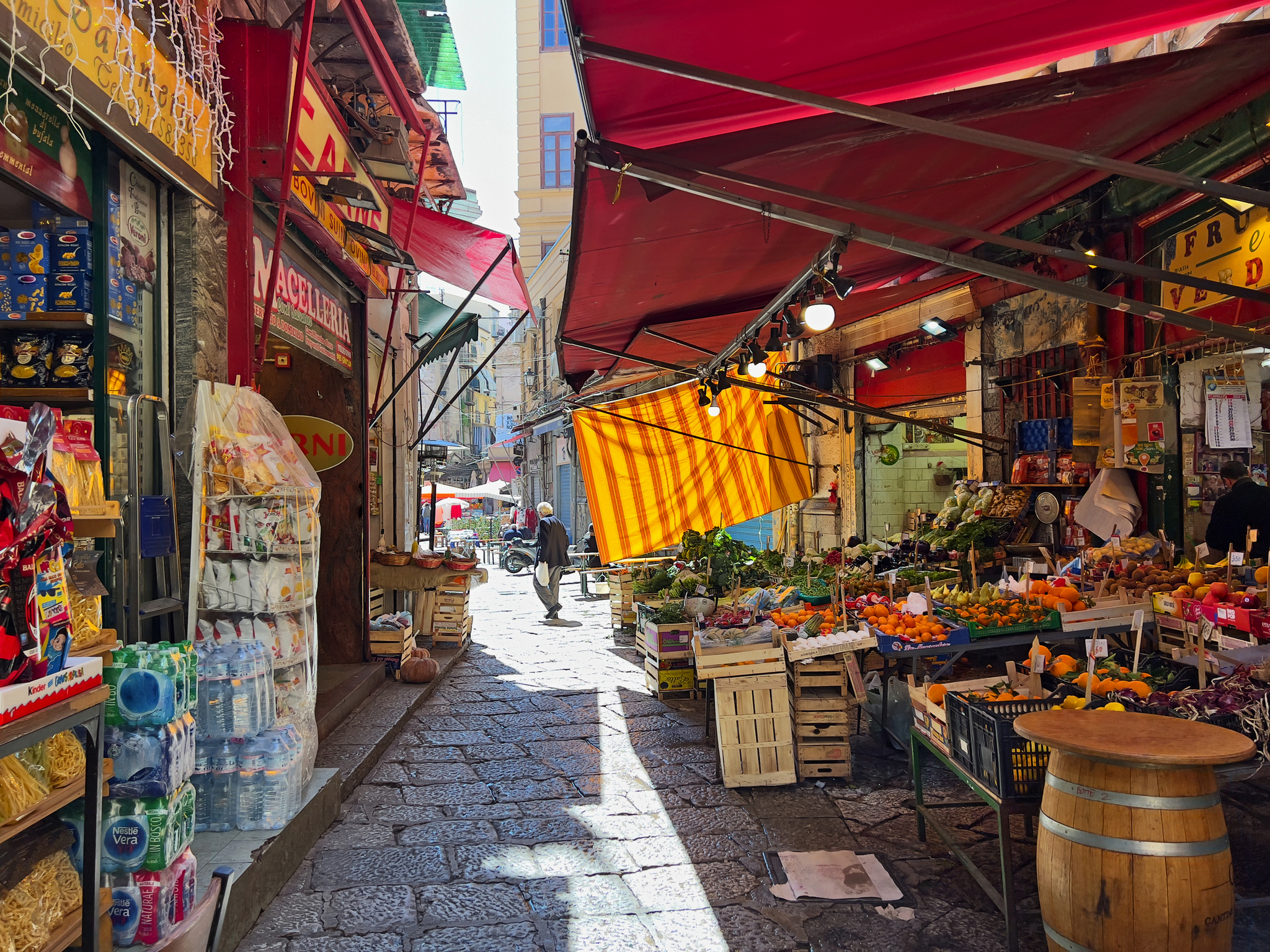 Marktstraße mit Obst- und Gemüseständen unter roten Sonnenschirmen.