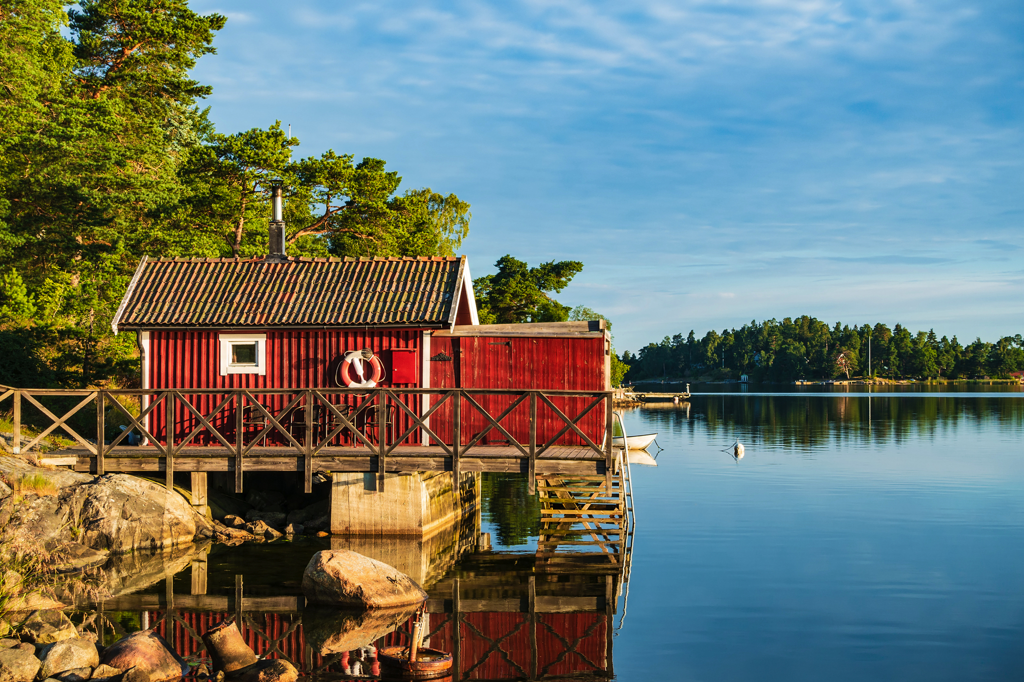 Rotes Holzhaus am Ufer eines Sees mit Bäumen im Hintergrund.