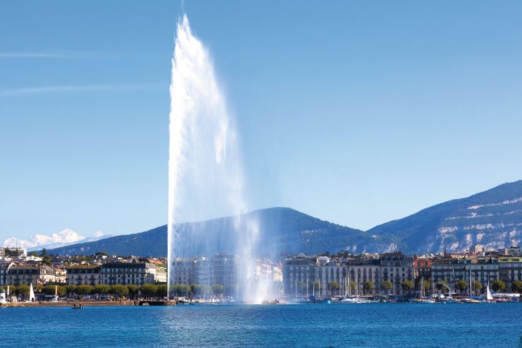 Großer Wasserfontäne am See mit Stadt und Bergen im Hintergrund.