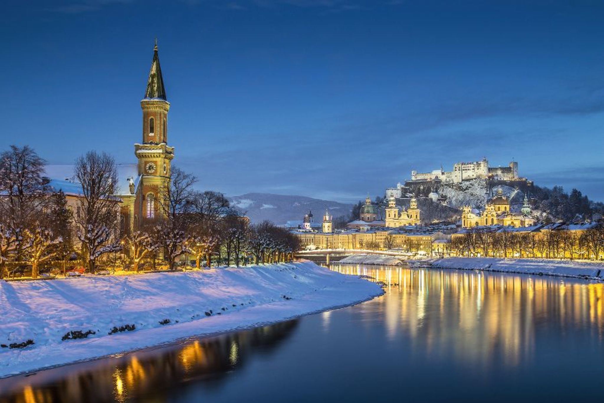 Winterliche Stadtansicht von Salzburg bei Abenddämmerung mit Schnee und beleuchtetem Fluss.