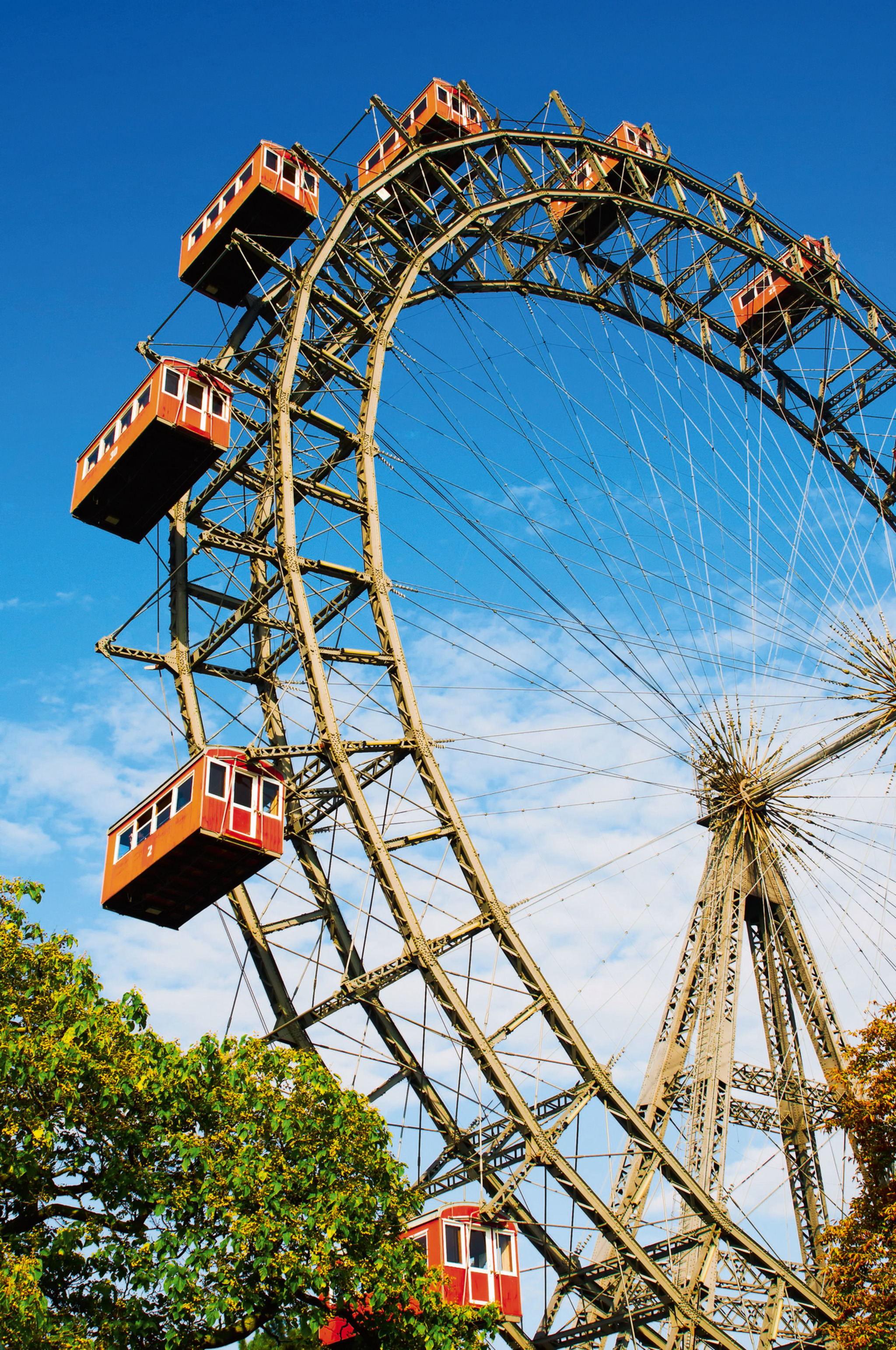Altes Riesenrad mit roten Gondeln vor blauem Himmel und grünen Bäumen.