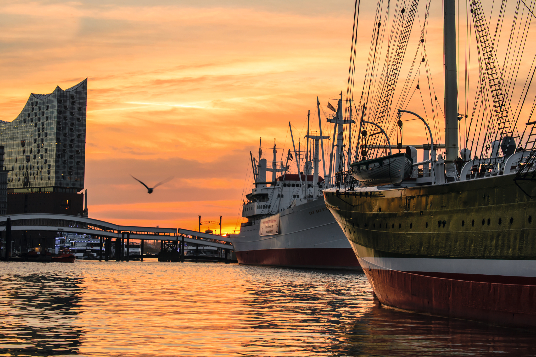 Schiffe im Hafen bei Sonnenuntergang, im Hintergrund moderne Architektur und ein Vogel.