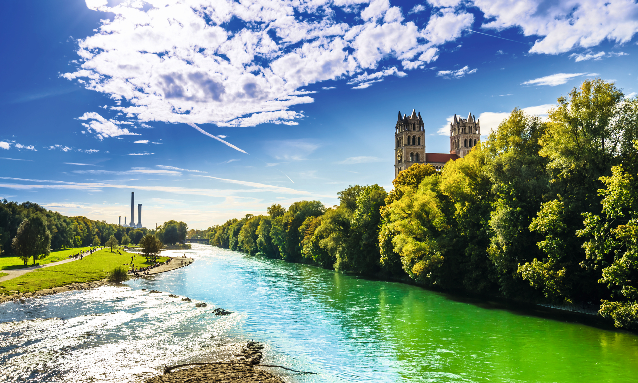 Fluss mit grünen Bäumen und einer Kathedrale im Hintergrund unter blauem Himmel.