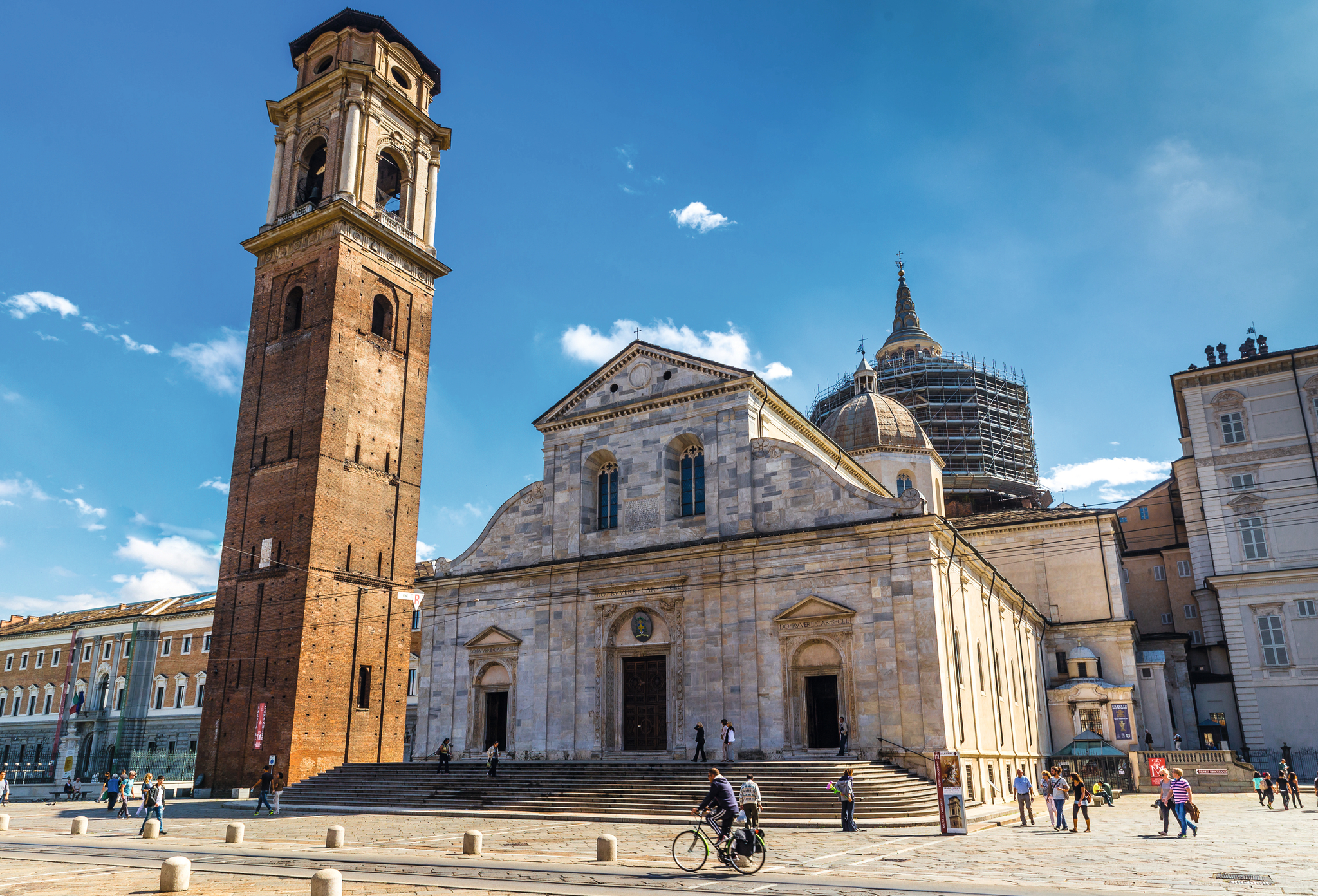 Kathedrale und Glockenturm auf einem sonnigen Platz, Menschen spazieren und radeln.
