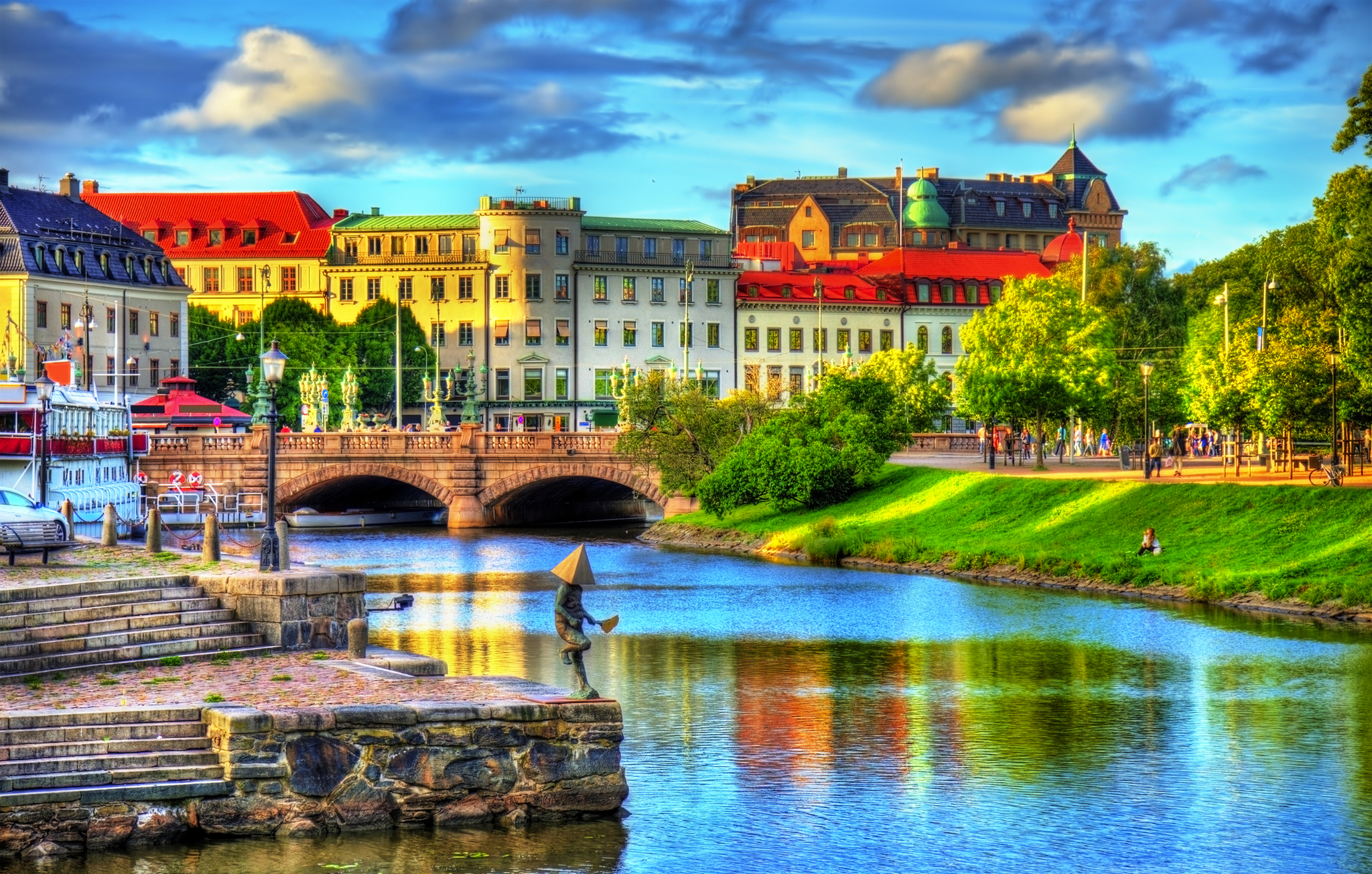 Blick auf eine malerische Stadtlandschaft mit einem Fluss im Vordergrund, kleinen Booten und einem steinernen Ufer. Im Hintergrund befinden sich historische Gebäude mit bunten Dächern und eine Brücke. Der Himmel ist blau mit einigen Wolken.