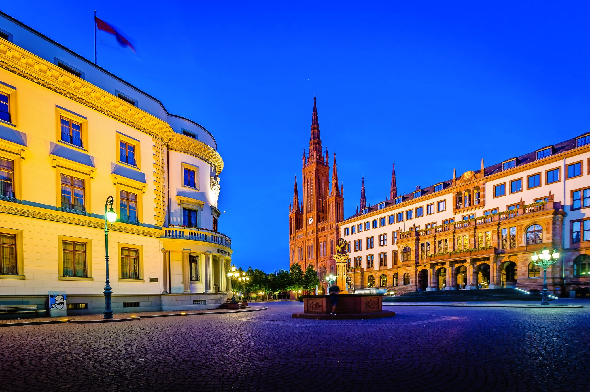 Wiesbaden Stadtplatz bei Nacht mit historischer Architektur, links ein beleuchtetes Gebäude mit Flagge, im Hintergrund eine gotische Kirche mit hohen Türmen, umgeben von weiteren historischen Gebäuden.