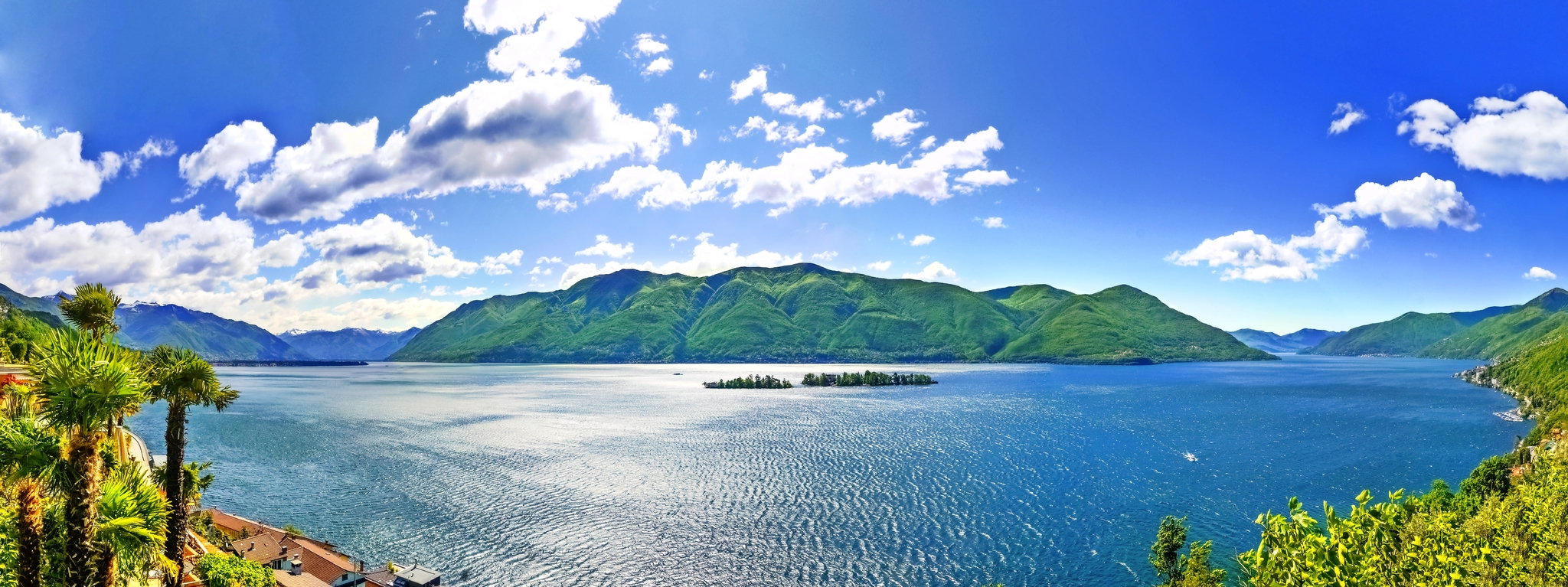 Blick auf einen See mit Inseln und bewaldeten Bergen im Hintergrund unter blauem Himmel.